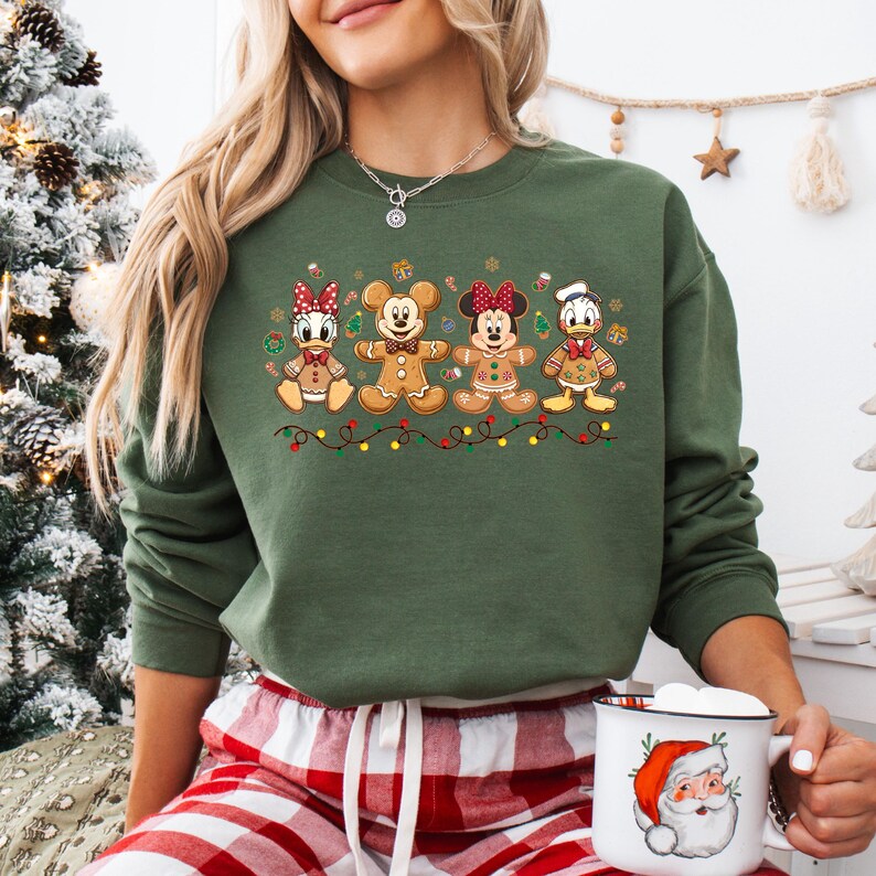 May include: A green sweatshirt featuring gingerbread versions of Disney characters: Daisy Duck, Mickey Mouse, Minnie Mouse, and Donald Duck. The design includes a string of lights and festive accents, perfect for the holiday season.
