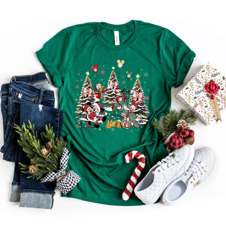 May include: Green t-shirt with a Christmas-themed design featuring Mickey Mouse, Minnie Mouse, Donald Duck, Goofy, and Pluto standing in front of decorated Christmas trees. The characters are wearing festive attire and holding presents.