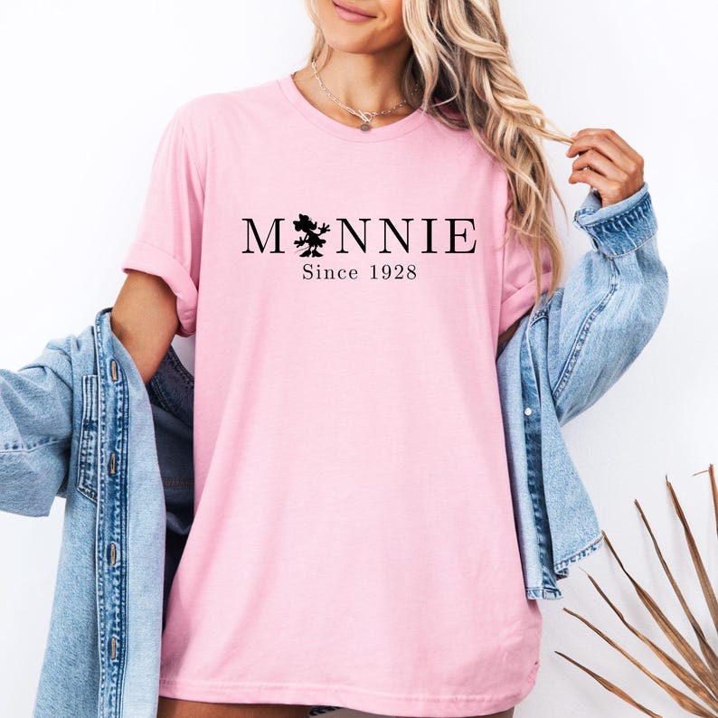 May include: A light pink t-shirt with the word "Minnie" in black, featuring a silhouette of Minnie Mouse. Below the word is "Since 1928". The shirt is worn with a denim jacket.