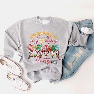 K&ouml;nnte beinhalten: Ein grauer Pullover mit einem Weihnachtsmotiv mit Mickey Mouse und seinen Freunden. Der Text auf dem Pullover lautet "Mickey's very merry Christmas Party 2025".