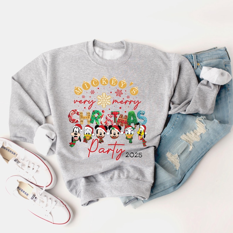 K&ouml;nnte beinhalten: Ein grauer Pullover mit einem Weihnachtsmotiv mit Mickey Mouse und seinen Freunden. Der Text auf dem Pullover lautet "Mickey's very merry Christmas Party 2025".