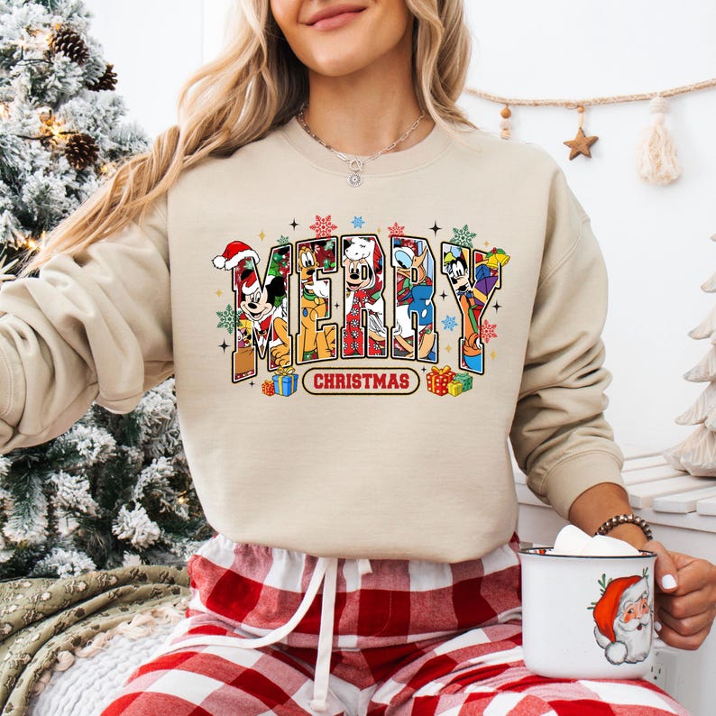 May include: A beige sweatshirt with the word "MERRY CHRISTMAS" in colorful block letters. The letters are filled with cartoon characters, snowflakes, and gift boxes. The sweatshirt is part of a holiday-themed collection.