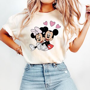 May include: A white t-shirt with a graphic of Mickey Mouse and Minnie Mouse hugging. The graphic is in black, red, and pink. There are three pink hearts above the graphic.