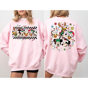 Puede incluir: Sudadera rosa de Navidad con un dise&ntilde;o festivo con Mickey Mouse, Minnie Mouse, Donald Duck, Goofy y Pluto. La parte delantera de la sudadera presenta a los personajes en fila con luces de Navidad y un patr&oacute;n a cuadros. La parte trasera de la sudadera presenta a los personajes en un c&iacute;rculo con luces de Navidad.