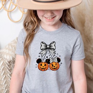 May include: A light gray t-shirt featuring a Halloween-themed design. The graphic includes two orange jack-o'-lanterns with mouse ears, a large bow, and a polka-dot bow. The design is centered on the shirt.