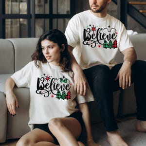 以下が含まれることがあります： 「Believe」と赤いチェック柄のクリスマスデザインがプリントされた白いTシャツを着たカップル。デザインには、城、星、ミッキーマウスの耳も含まれています。