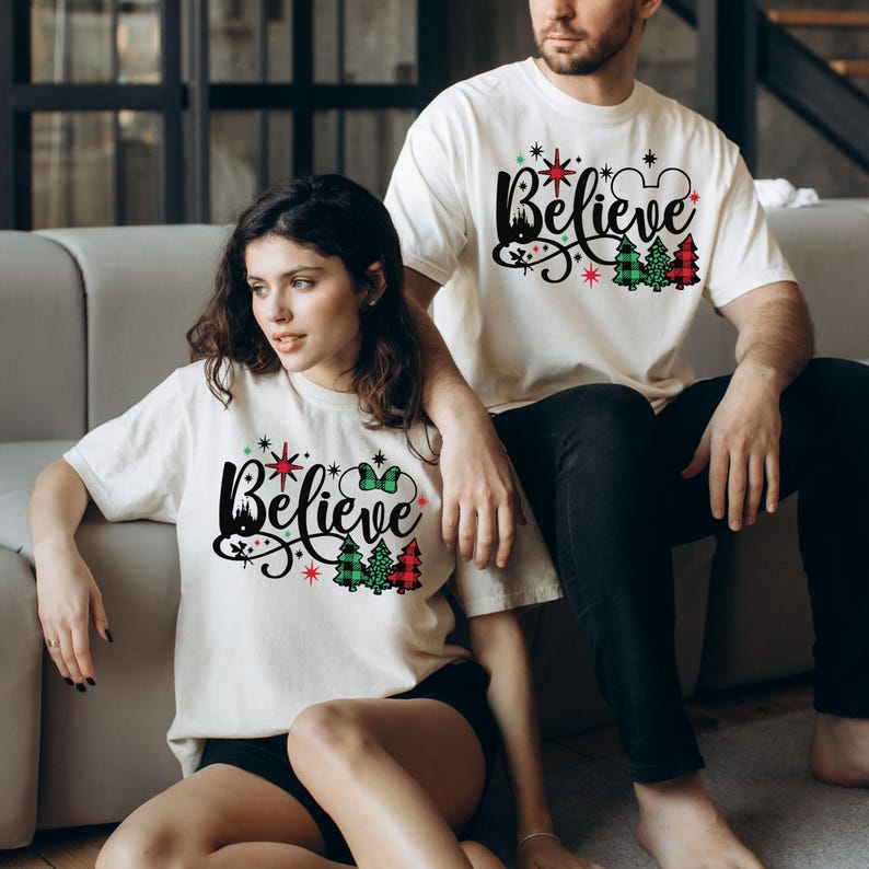 以下が含まれることがあります： 「Believe」と赤いチェック柄のクリスマスデザインがプリントされた白いTシャツを着たカップル。デザインには、城、星、ミッキーマウスの耳も含まれています。