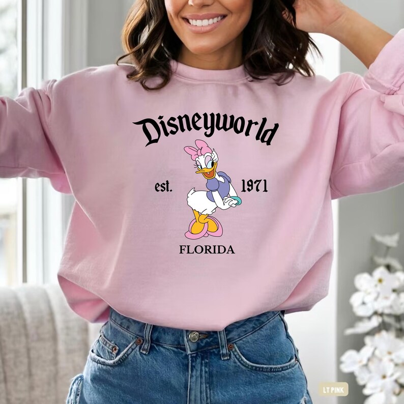 May include: A light pink sweatshirt featuring the text "Disneyworld" in black script above a graphic of Daisy Duck. The graphic includes the text "est. 1971" and "FLORIDA".
