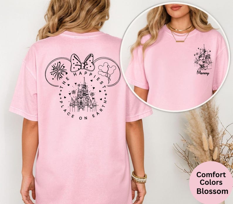 May include: A light pink t-shirt with a black graphic on the back featuring Minnie Mouse ears, a bow, fireworks, balloons, a castle, and the text "THE HAPPIEST PLACE ON EARTH." The front of the shirt has a smaller graphic with a castle and the word "Mommy."