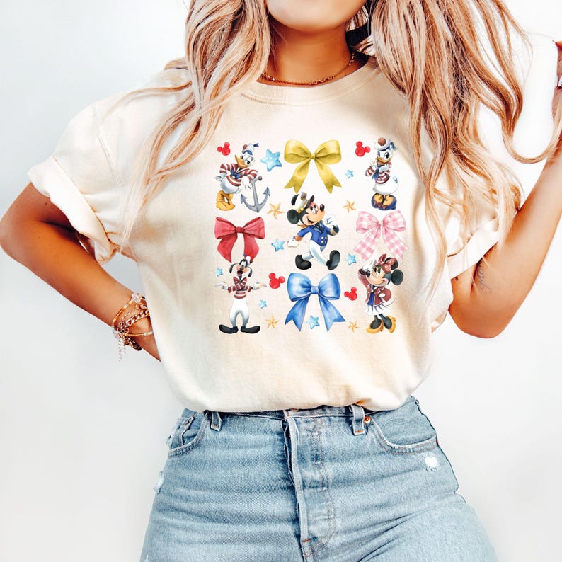 May include: A white t-shirt with a colorful graphic print of Mickey Mouse and friends wearing sailor outfits and holding anchors and stars. The graphic also includes red, blue, and yellow bows.
