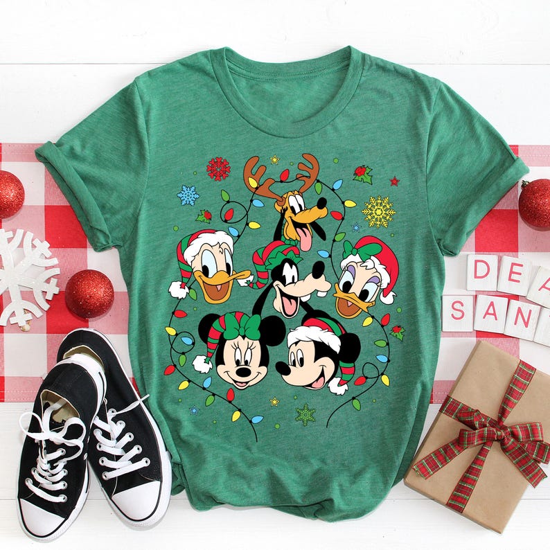 May include: Green t-shirt with a Christmas-themed design featuring Mickey Mouse, Minnie Mouse, Donald Duck, Goofy, and Pluto. The characters are surrounded by Christmas lights and snowflakes.