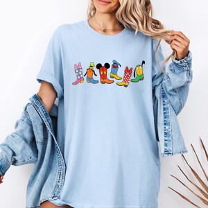 May include: Light blue t-shirt featuring a colorful graphic of cartoon cowboy boots. The boots are in various colors and patterns, including pink, orange, red, yellow, and green, with character-inspired designs. The shirt has a relaxed fit.