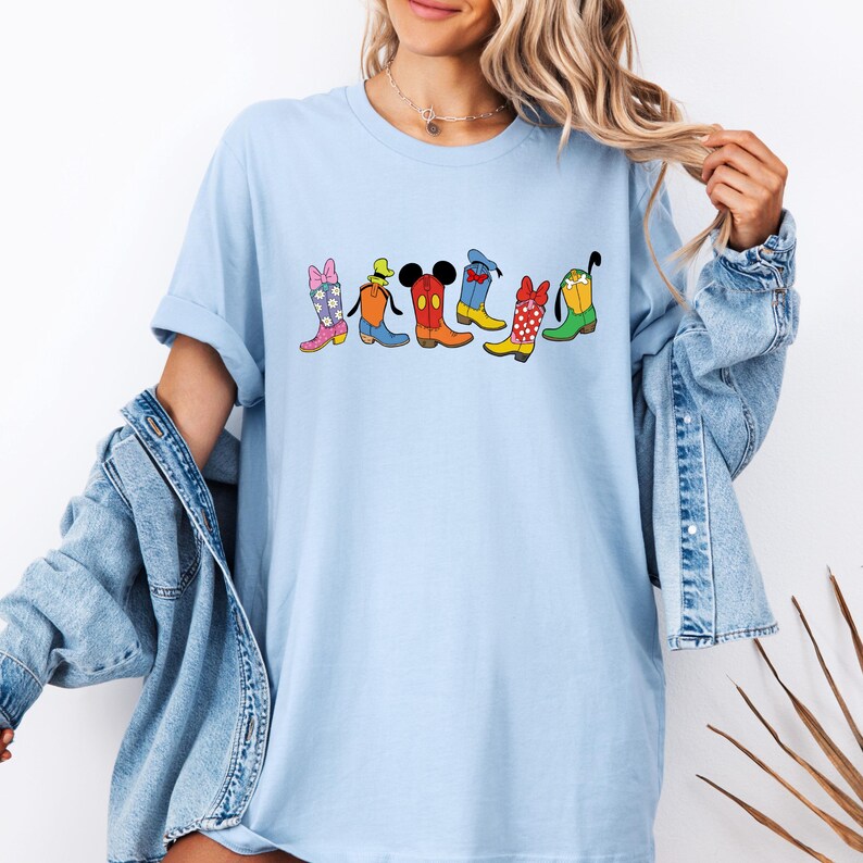 May include: Light blue t-shirt featuring a colorful graphic of cartoon cowboy boots. The boots are in various colors and patterns, including pink, orange, red, yellow, and green, with character-inspired designs. The shirt has a relaxed fit.