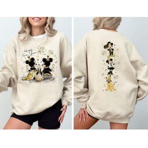 Puede incluir: Sudadera beige con Mickey y Minnie Mouse celebrando el A&ntilde;o Nuevo. La parte delantera muestra el texto "Happy New Year" con fuegos artificiales y los personajes. La parte trasera muestra a Mickey, Minnie y Pluto con ropa festiva.