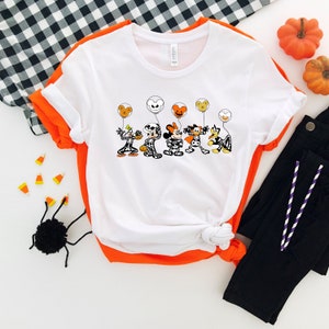 Op de afbeelding: Wit T-shirt met een afbeelding van Mickey Mouse, Minnie Mouse, Donald Duck, Goofy en Pluto gekleed in skeletkostuums en oranje en gele ballonnen vasthoudend. De personages lachen allemaal en hebben zwarte ogen. Het shirt ligt op een witte achtergrond met een zwart-wit geruit patroon.