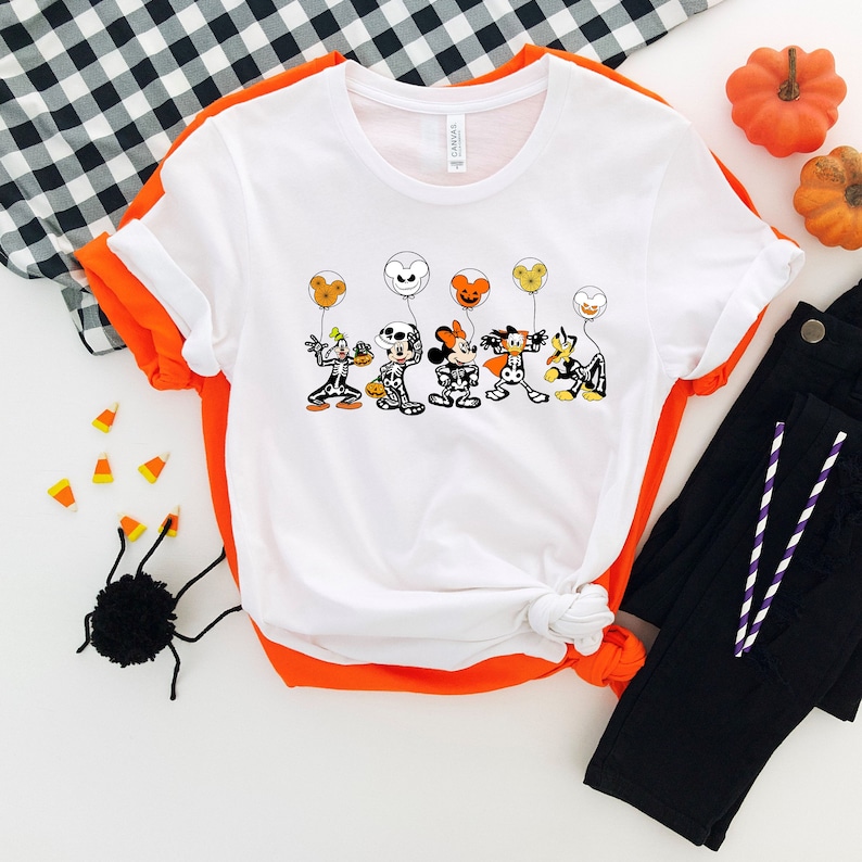 Op de afbeelding: Wit T-shirt met een afbeelding van Mickey Mouse, Minnie Mouse, Donald Duck, Goofy en Pluto gekleed in skeletkostuums en oranje en gele ballonnen vasthoudend. De personages lachen allemaal en hebben zwarte ogen. Het shirt ligt op een witte achtergrond met een zwart-wit geruit patroon.