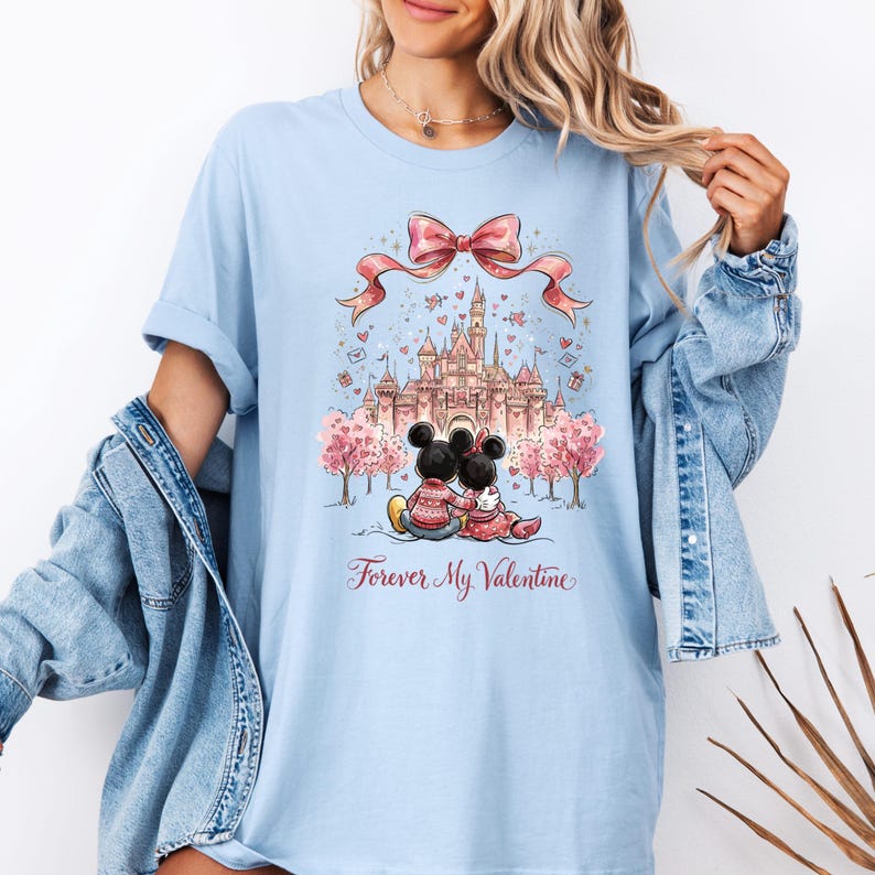May include: Light blue t-shirt featuring a graphic of Mickey and Minnie Mouse embracing in front of a castle, with a pink bow and the text "Forever My Valentine." The design includes pink trees and hearts.