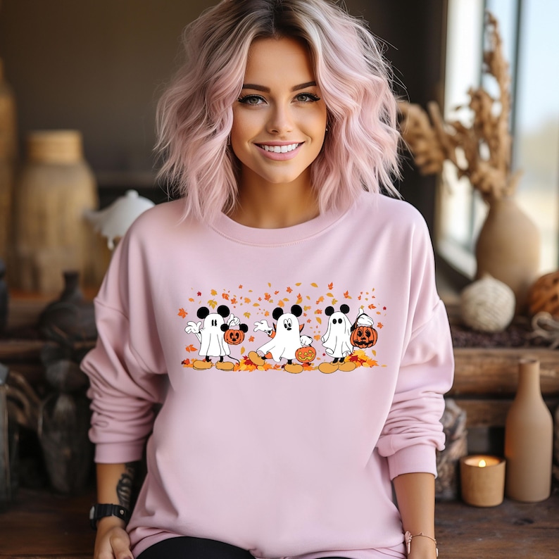 May include: Pink long-sleeved sweatshirt with a graphic of three Mickey Mouse characters dressed as ghosts carrying jack-o'-lanterns. The characters are walking in a line with autumn leaves falling around them.