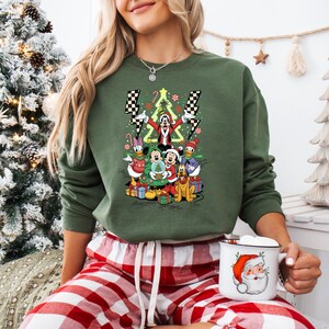 May include: Green crewneck sweatshirt featuring a festive Disney character design with Mickey, Minnie, Donald, and Goofy. The design includes a Christmas tree, presents, and lightning bolts. The person is wearing red and white plaid pajama pants and holding a Santa mug.