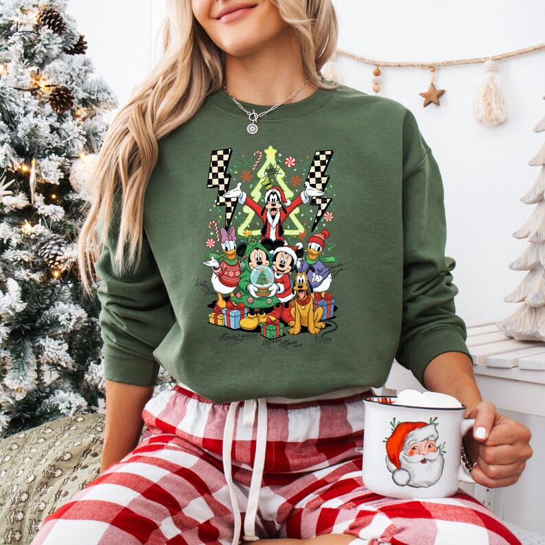 May include: Green crewneck sweatshirt featuring a festive Disney character design with Mickey, Minnie, Donald, and Goofy. The design includes a Christmas tree, presents, and lightning bolts. The person is wearing red and white plaid pajama pants and holding a Santa mug.