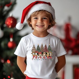 May include: A child wearing a white t-shirt featuring a Christmas design with cartoon characters and Christmas trees. The child is also wearing a red Santa hat with a white pom-pom. The background includes a Christmas tree with red ornaments.