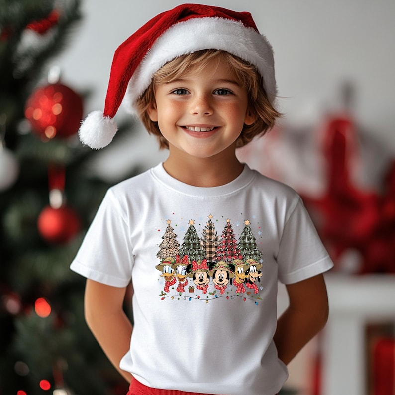 May include: A child wearing a white t-shirt featuring a Christmas design with cartoon characters and Christmas trees. The child is also wearing a red Santa hat with a white pom-pom. The background includes a Christmas tree with red ornaments.