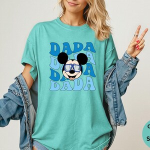 K&ouml;nnte beinhalten: T&uuml;rkisfarbenes T-Shirt mit dem Wort "DADA" in blauer Retro-Schrift, dreimal wiederholt. Eine Zeichentrickmaus mit blauer Brille ist unter dem Text zentriert. Das Shirt geh&ouml;rt zur Comfort Colors Kollektion.