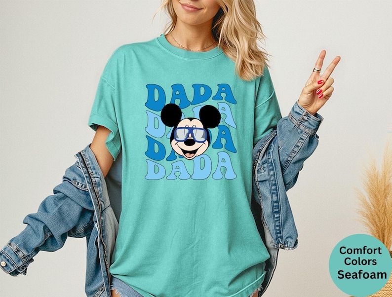 K&ouml;nnte beinhalten: T&uuml;rkisfarbenes T-Shirt mit dem Wort "DADA" in blauer Retro-Schrift, dreimal wiederholt. Eine Zeichentrickmaus mit blauer Brille ist unter dem Text zentriert. Das Shirt geh&ouml;rt zur Comfort Colors Kollektion.