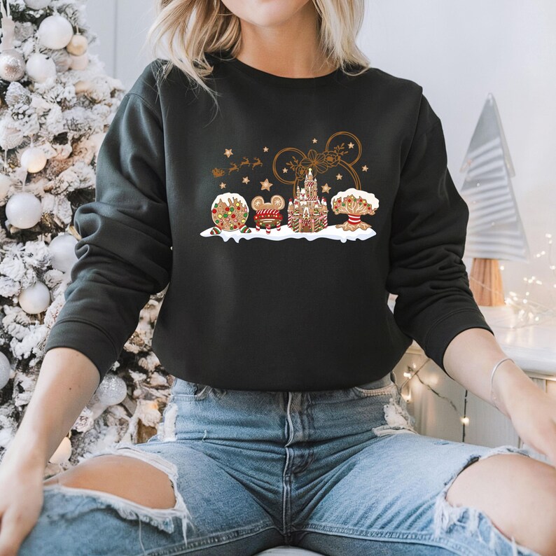 May include: Black sweatshirt featuring a festive gingerbread design. The design includes a gingerbread Mickey Mouse head, castle, and other holiday elements. The sweatshirt is worn by a person wearing blue jeans.