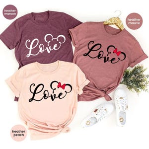 K&ouml;nnte beinhalten: Drei T-Shirts mit dem Wort "Love" in einer Schriftart mit einer Mickey-Maus-Silhouette und einer roten Schleife. Die Shirts sind in Heather Maroon, Heather Mauve und Heather Peach.
