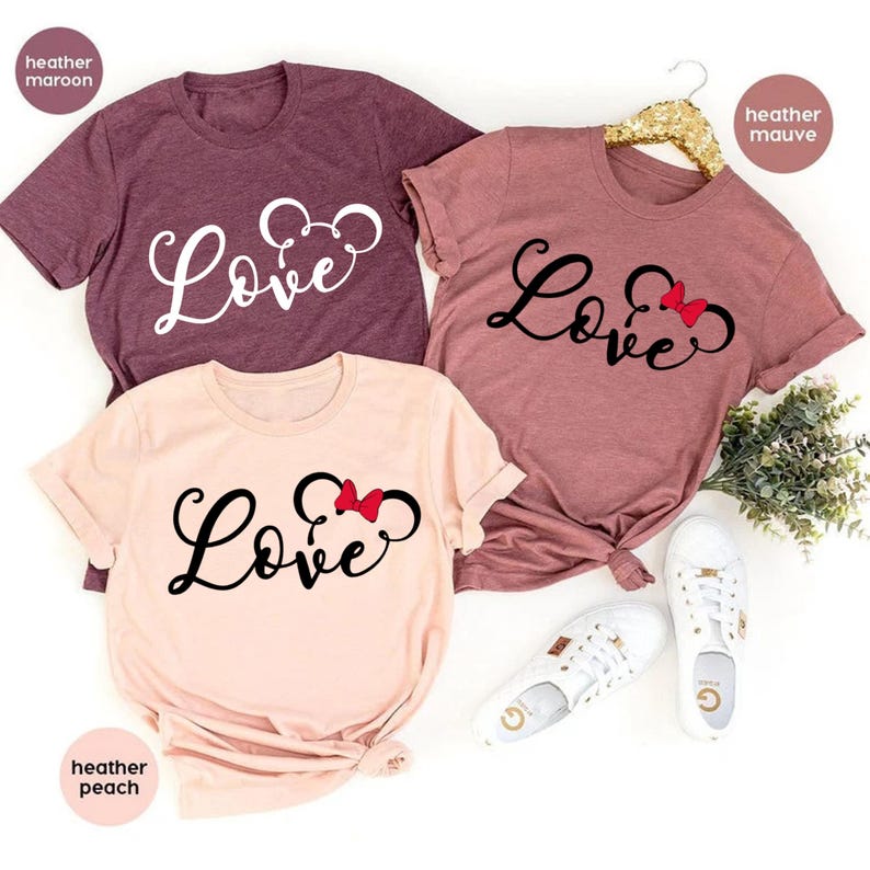 K&ouml;nnte beinhalten: Drei T-Shirts mit dem Wort "Love" in einer Schriftart mit einer Mickey-Maus-Silhouette und einer roten Schleife. Die Shirts sind in Heather Maroon, Heather Mauve und Heather Peach.