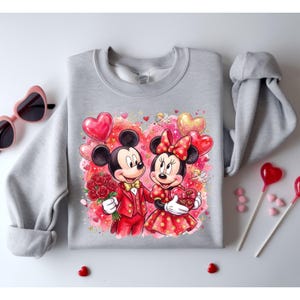 May include: A gray sweatshirt featuring a Valentine's Day design with Mickey and Minnie Mouse. Mickey is holding roses and Minnie holds a heart-shaped box of chocolates. The background includes heart-shaped balloons and cherubs.