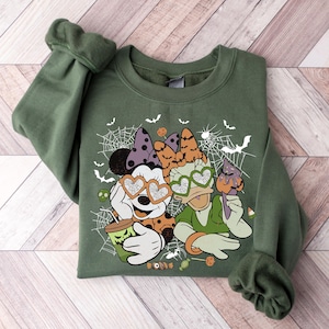 May include: A green sweatshirt with a Halloween-themed design featuring Mickey Mouse and Minnie Mouse wearing heart-shaped sunglasses. The design includes spiderwebs, bats, and candy.