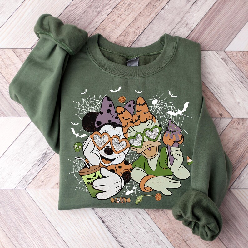 May include: A green sweatshirt with a Halloween-themed design featuring Mickey Mouse and Minnie Mouse wearing heart-shaped sunglasses. The design includes spiderwebs, bats, and candy.