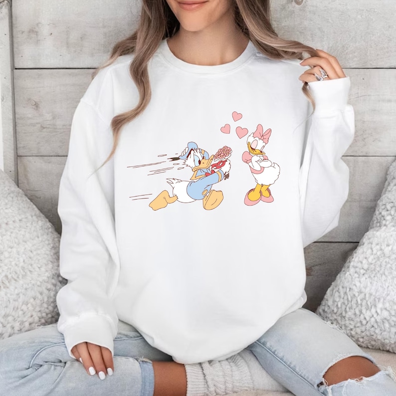 Op de afbeelding: Wit sweatshirt met een cartoon afbeelding van Donald Duck die met een bos bloemen naar Daisy Duck rent, die met gekruiste armen en een roze strik in haar haar staat. Er zweven roze hartjes tussen hen.