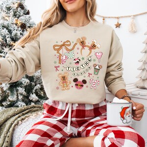 May include: A beige sweatshirt with a festive graphic featuring gingerbread men, candy canes, and the text "Tis the Season." The design includes Mickey Mouse-inspired elements and holiday treats. The sweatshirt is paired with red and white plaid pajama bottoms.