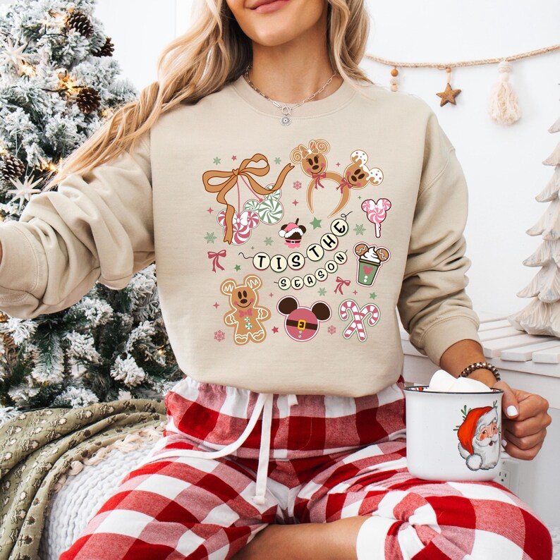 May include: A beige sweatshirt with a festive graphic featuring gingerbread men, candy canes, and the text "Tis the Season." The design includes Mickey Mouse-inspired elements and holiday treats. The sweatshirt is paired with red and white plaid pajama bottoms.