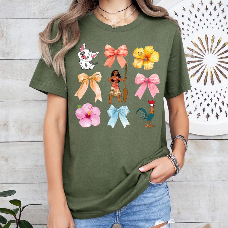Pu&ograve; includere: Una maglietta verde con una variet&agrave; di immagini colorate, tra cui un gallo, un fiore di ibisco, un fiocco e un personaggio dei cartoni animati. Il personaggio dei cartoni animati &egrave; una donna polinesiana con i capelli castani e un vestito marrone. Tiene una pagaia e sta in piedi su una spiaggia.