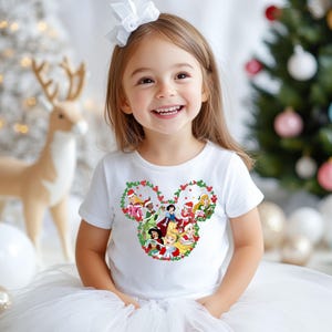 Pu&ograve; includere: Una bambina sorride, indossa una maglietta bianca con un design festivo a forma di testa di Topolino. Il design &egrave; pieno di principesse Disney in abiti natalizi, circondate da accenti rossi e verdi. Lo sfondo include un albero di Natale e decorazioni a forma di renna.