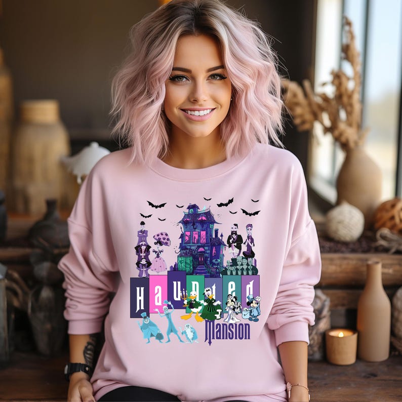 Puede incluir: Una sudadera rosa con un estampado gr&aacute;fico de la Mansi&oacute;n Encantada de Disneyland. El gr&aacute;fico presenta la mansi&oacute;n, fantasmas y el texto "Haunted Mansion".