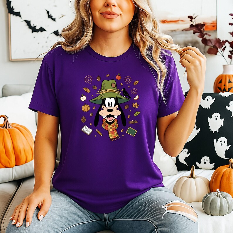 Puede incluir: Una camiseta morada con un personaje de dibujos animados que lleva un sombrero verde y una bufanda, rodeado de ilustraciones con tem&aacute;tica oto&ntilde;al. El dise&ntilde;o incluye calabazas, manzanas y hojas.