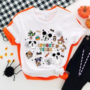 May include: White t-shirt featuring a colourful graphic of various Disney characters in Halloween costumes. The text "Spooky Vibes" is displayed in the centre of the design.