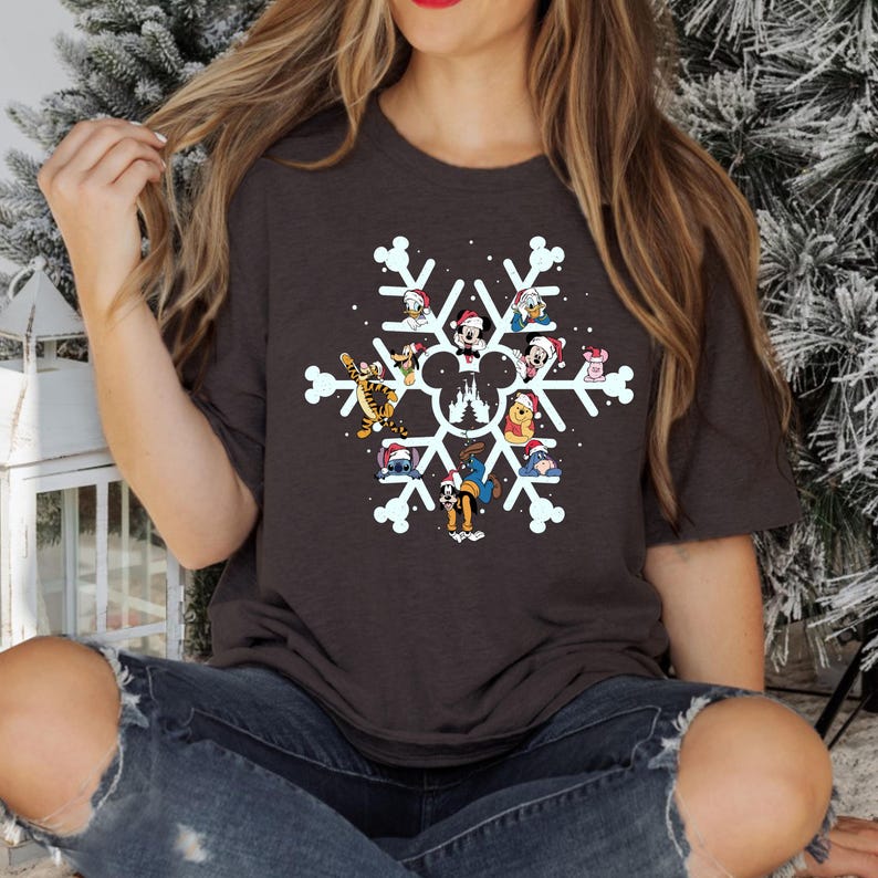 May include: A dark gray t-shirt with a white snowflake design featuring various Disney characters, including Mickey Mouse, Donald Duck, Goofy, and others. The characters are wearing Santa hats and are arranged around a white silhouette of the Disney castle.