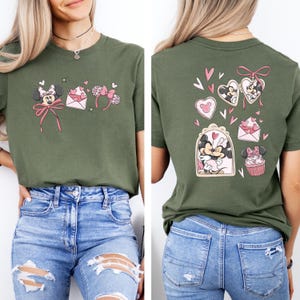May include: Olive green t-shirt featuring Minnie Mouse-themed graphics on the front and back. The front design includes a bow, ears, and an envelope with hearts. The back design showcases Minnie and Mickey Mouse in heart-shaped frames and a cupcake.