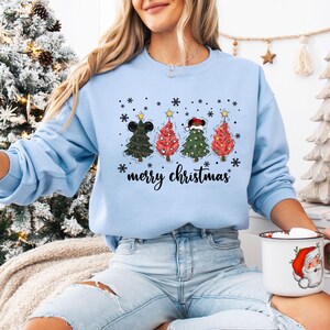 May include: Light blue sweatshirt with a graphic of four Christmas trees, each adorned with a different hat and star. The text "merry christmas" is written below the trees. Snowflakes are scattered around the trees.