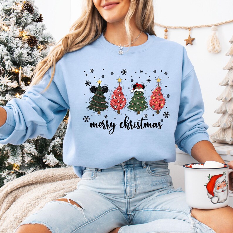May include: Light blue sweatshirt with a graphic of four Christmas trees, each adorned with a different hat and star. The text "merry christmas" is written below the trees. Snowflakes are scattered around the trees.