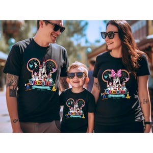 May include: A family of three wearing black t-shirts with a Disneyland graphic featuring Mickey Mouse, Minnie Mouse, and the Disneyland castle. The graphic also includes fireworks and the text "Disneyland" and "The happiest place on earth est. 1955."