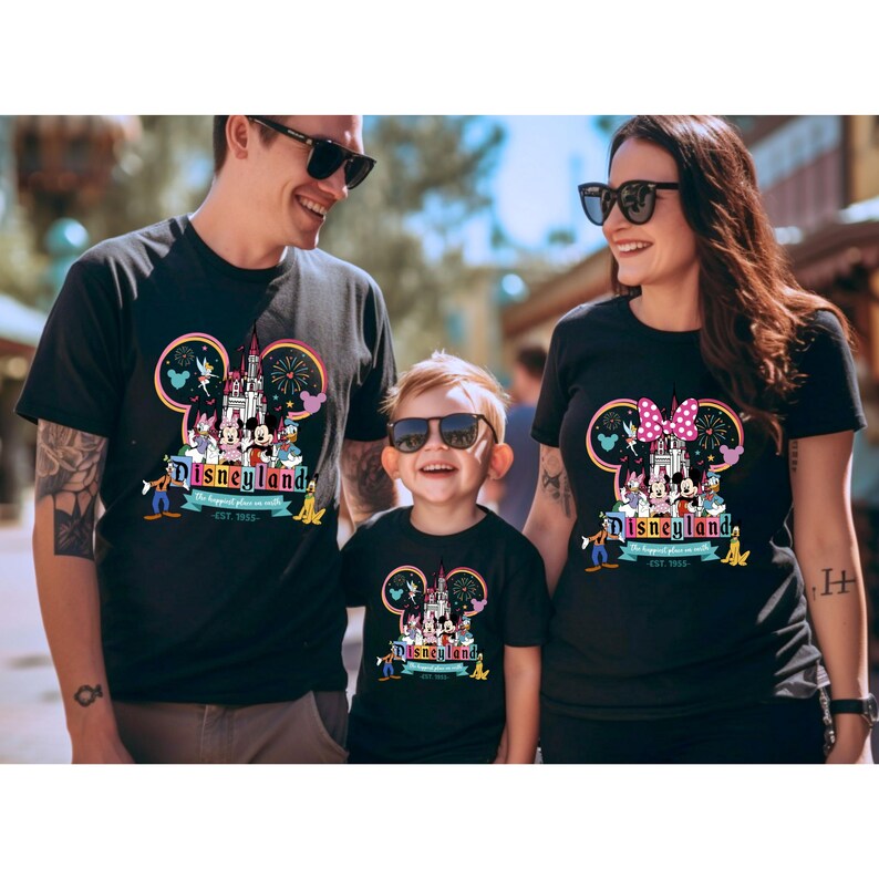 May include: A family of three wearing black t-shirts with a Disneyland graphic featuring Mickey Mouse, Minnie Mouse, and the Disneyland castle. The graphic also includes fireworks and the text "Disneyland" and "The happiest place on earth est. 1955."