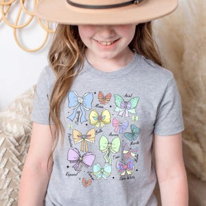 May include: A gray t-shirt with a colorful design of bows and butterflies. Each bow is labeled with the name of a Disney princess: Ariel, Moana, Cinderella, Belle, Aurora, Merida, Tiana, Mulan, Rapunzel, Jasmine, and Snow White.