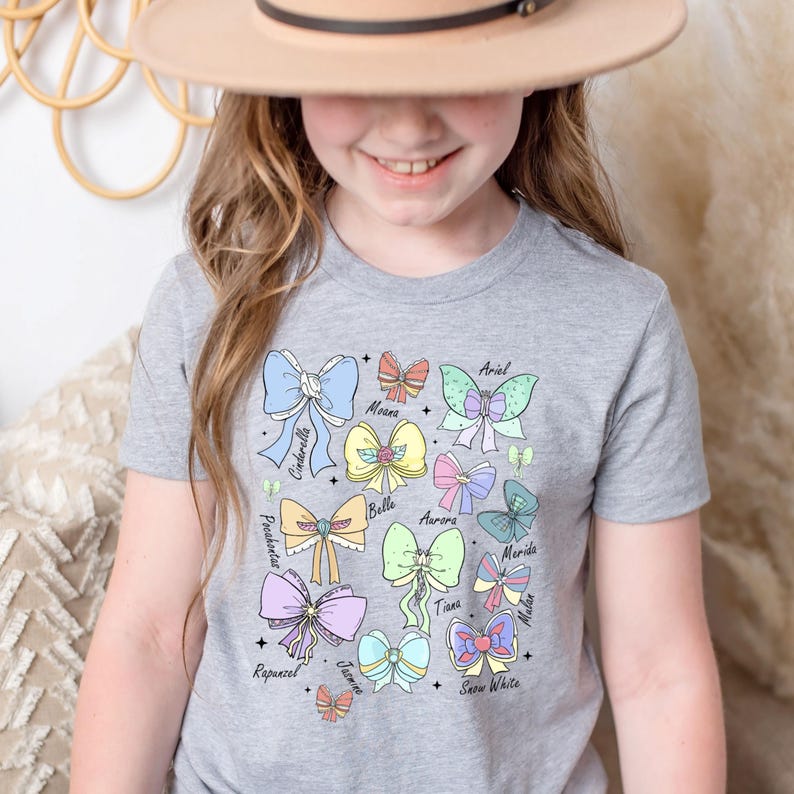 May include: A gray t-shirt with a colorful design of bows and butterflies. Each bow is labeled with the name of a Disney princess: Ariel, Moana, Cinderella, Belle, Aurora, Merida, Tiana, Mulan, Rapunzel, Jasmine, and Snow White.